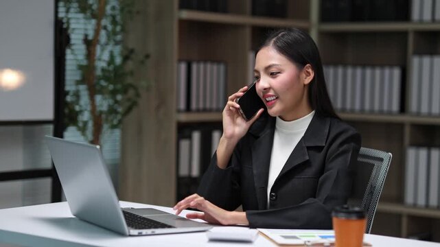 Professional asian businesswoman working on laptop while talking on mobile phone in a modern office with a positive attitude