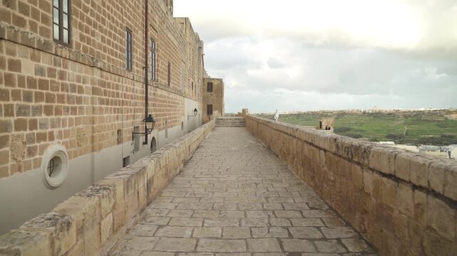 Walking along ancient stone fortress rampart overlooking rolling green hills and distant town.