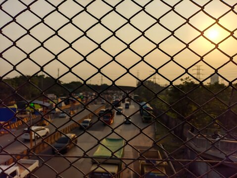 Highway traffic view in the evening through a metal chain link wire fence with bright sunset flare