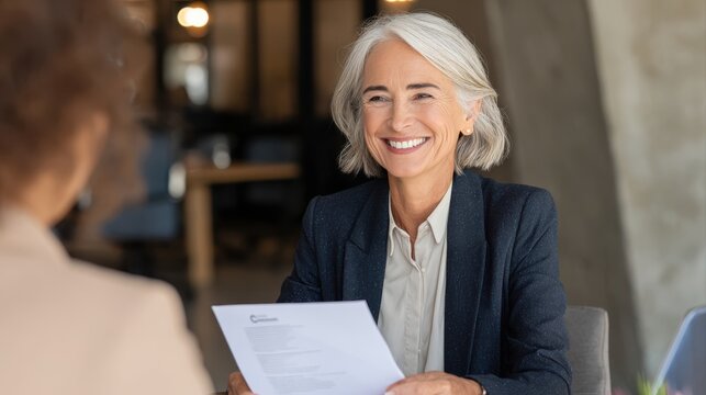 Smiling mature business woman hr holding cv document at job interview. Happy mid aged professional banking manager or lawyer consulting client sitting at workplace in corporate office meeting.