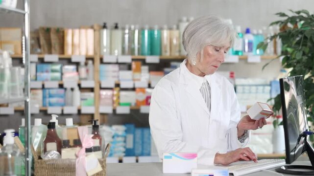 Mature female pharmacist standing behind the counter and working on computer, she checks the availability of medical medicines in a spreadsheet and prepares to collect an online order . High quality