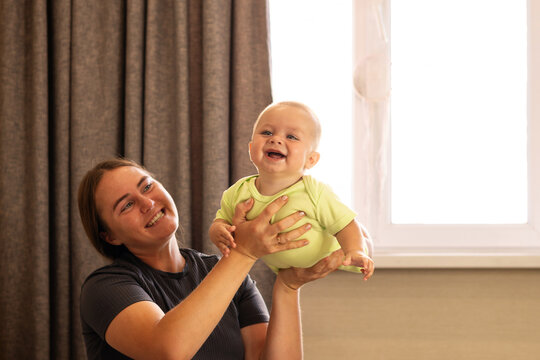 Happy Mother Lifts Laughing Baby Son Near Window