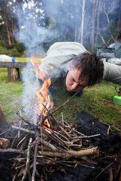 A young man in his twenties blowing on a small campfire to help it keep burning