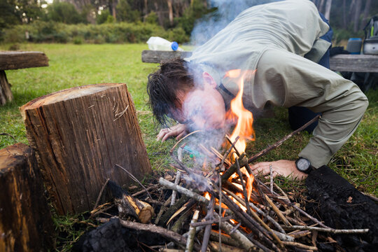 A young man in his twenties blowing on a small campfire to help it keep burning