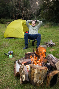 Young man relaxing sitting in front of a campfire and yellow coloured tent in background