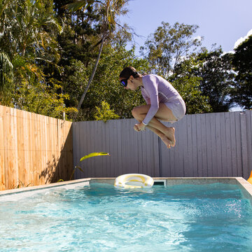 Young girl mid‑air above a backyard pool with knees tucked as she prepares to make a splash