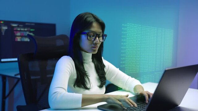 Young woman software developer types code on a laptop in a dark blue office at night, with green digital code floating beside her and data dashboards on monitors behind.