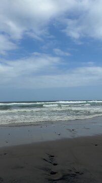 Scenic vertical view of ocean waves rolling onto a sandy beach under a blue sky with soft clouds.