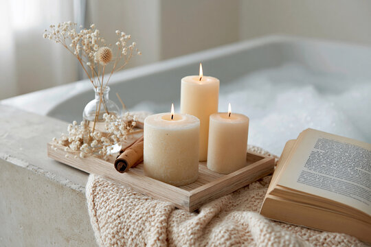 Soft candlelight glows warmly beside a bubbling bathtub. Candles rest on a wooden tray with dried flowers and a cinnamon stick. An open book lies nearby, inviting quiet reading