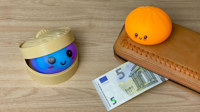 An aerial view of two differently coloured dumpling shaped squishies next to a leather wallet and a 5 euro banknote against a wooden surface.
