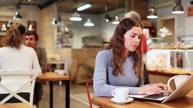 In cozy cafe hall, freelance girl works with laptop and drinks coffee. Remote work from anywhere in world 