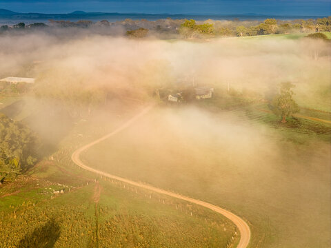 Aerial view of a winding dirt road curves through green fields leading to a farmhouse