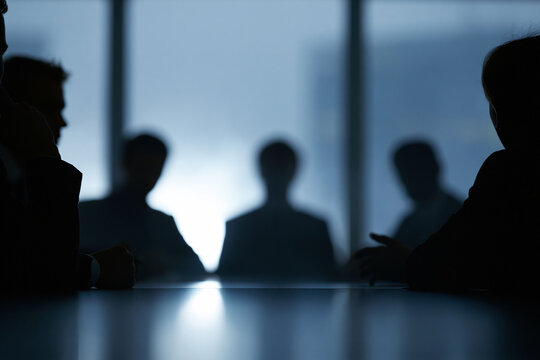 Silhouettes gather around a polished table. Soft light glows behind them, blurred and bright. Expressions hidden, attention focused on discussion. Modern office or conference room setting implied
