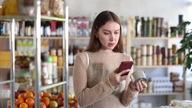 Girl customer at shop scan QR code and take reading on tin cans of preserves using phone camera. Client study composition, manufacturer information of product using application. Visitor examine