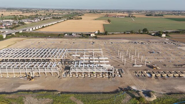 Logistics park construction in Barabasca near the Fiorenzuola d'Arda A1 motorway exit, with steel warehouse frames, construction yards and cultivated Po Valley farmland, drone slide