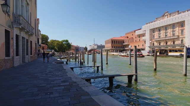 A moving view along the paved quayside where pedestrians pass shuttered palazzi, with mooring piles and a submerged plank in the green water alongside, in Venice, Italy.