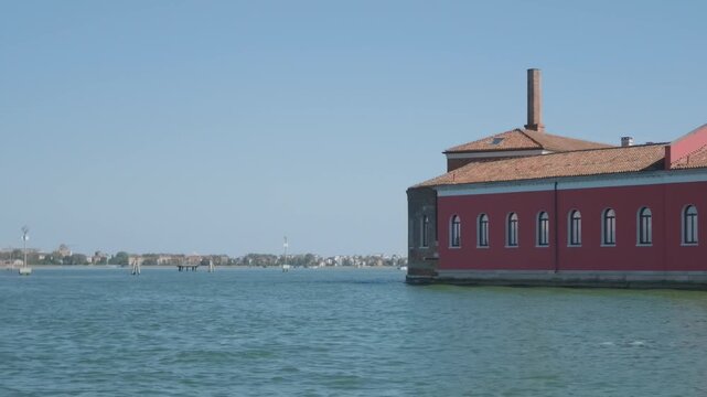 A deep red rectangular block with arched windows and a tall brick chimney occupies a low island, with the distant shoreline visible beyond, in Venice, Italy.