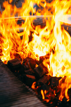 Flames dance above glowing charcoal in a grill during an outdoor cooking event. The bright fire contrasts with the dark coals, creating a lively scene