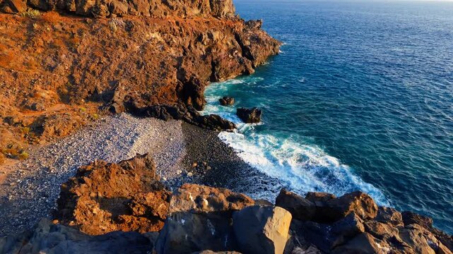 Waves crash on a rocky beach below steep cliffs in warm evening sunlight