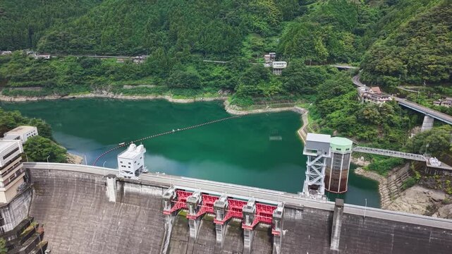 High-angle drone view showing the top structure of a concrete dam wall and green reservoir water in mountain wilderness.