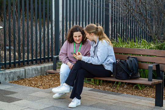 Two young women engaged in conversation while sitting on an urban bench looking at a smartphone