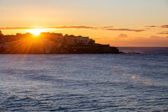 Golden sunset over ocean beside a densely developed coastal headland with dramatic sun rays