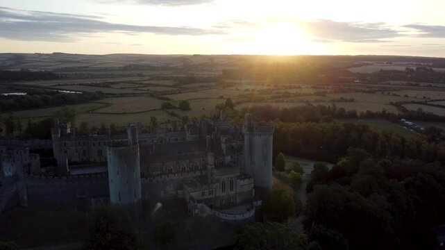 Cinematic wide drone shot from the side of Arundel Castle at sunrise, slowly panning towards the rear to showcase its historic silhouette against the glowing West Sussex landscape