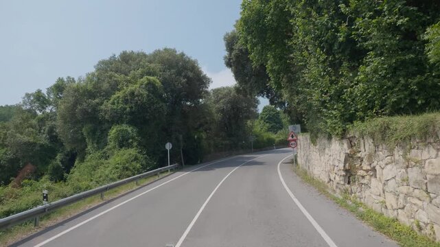 Country road curving through green trees, passing a stone wall and traffic signs, suggesting travel and journey