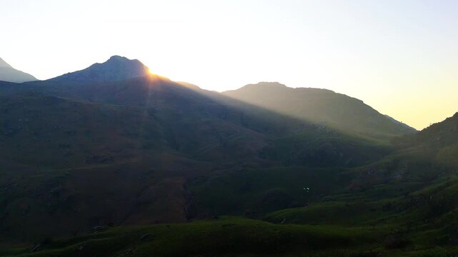 Drone tracks laterally as the setting sun emerges behind a mountain ridge, casting warm golden rays across the green slopes of Mulanje in Malawi.