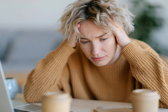 Young woman sits at desk, hands on head. She looks tired, frustrated, or overwhelmed. Coffee cups rest beside her laptop. Soft light illuminates her thoughtful expression. Perfect for scenes of stress