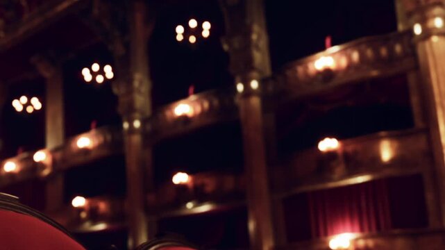 The inside of an old theater during a performance. Lights glow in the background, illuminating the rich design and structure. Red curtains add depth to the setting.