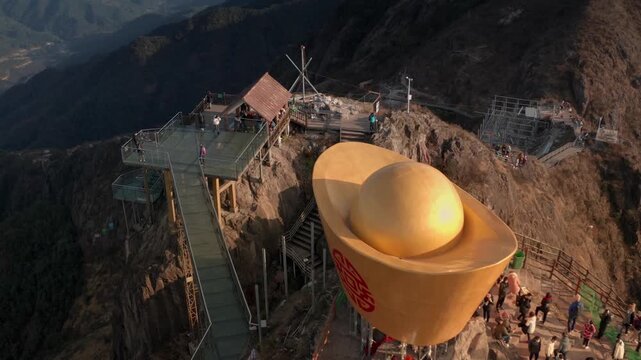 Aerial motion of people on a mountain peak with a giant golden ingot statue and scenic mountain range background.