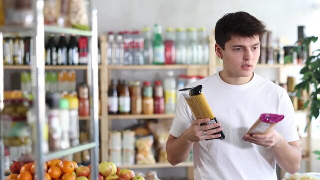 Positive young guy standing in grocery store aisle and holding two packs of spaghetti, carefully considering choice, comparing prices, ingredients, and quality before buying . High quality 4k footage