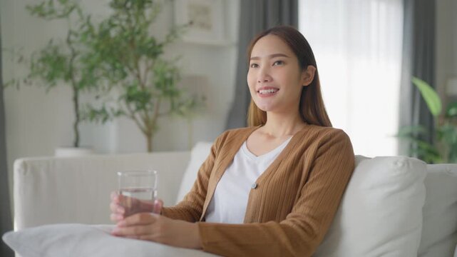 Happy young Asian woman drinking water while sitting on the sofa in the living room