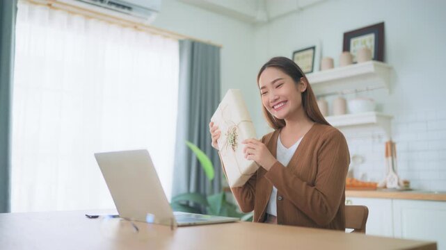 Young asian woman using computer laptop at home. Female showing gift box while on video conversation with friend. Happy birthday, Happy new year, Thanksgiving day