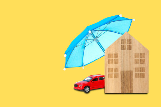 Insurance. Wooden house and red car placed under umbrella on yellow backdrop