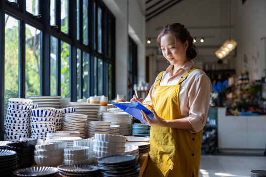 Asian employee is examine her new ceramics shelf of bowl, plate, cutlery and utensil in local artisan handmade store for handcraft and products for part time job concept