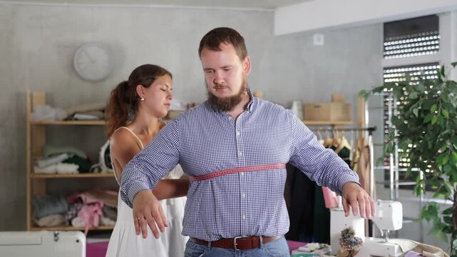 Young seamstress skillfully taking measurements for male client in modest sewing studio, preparing for custom-made clothing order 