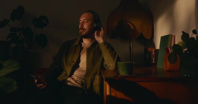 A bearded man sits at a cozy, dimly lit desk at home with eyes closed, listening to audio on headphones while holding his phone, enjoying a warm evening with a coffee mug, plants, and lamplight.