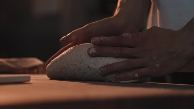 Male baker hands shaping fresh bread dough on rustic wooden table, preparing artisanal bread loaves and rolls in cozy small bakery kitchen, warm lighting ambiance