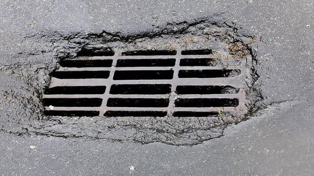 Drain grate on wet asphalt road surface with water flowing into opening, pavement showing signs of wear and tear, close up view of street drain cover