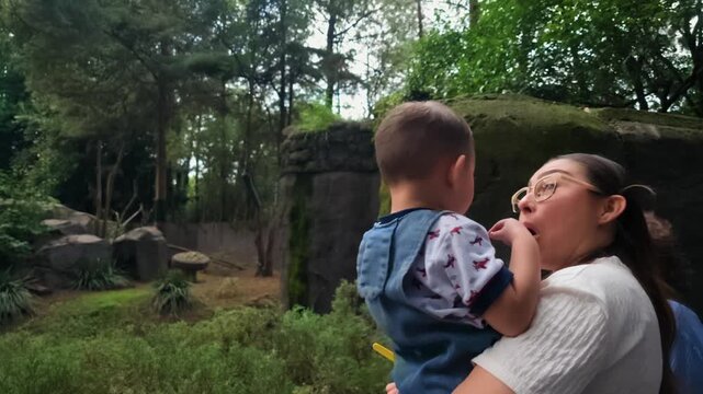 A loving Latina mother and grandmother hold a happy 2-year-old baby, observing the brown bear habitat at Chapultepec Zoo, engaging in playful singing and dancing. This heartwarming family moment captu