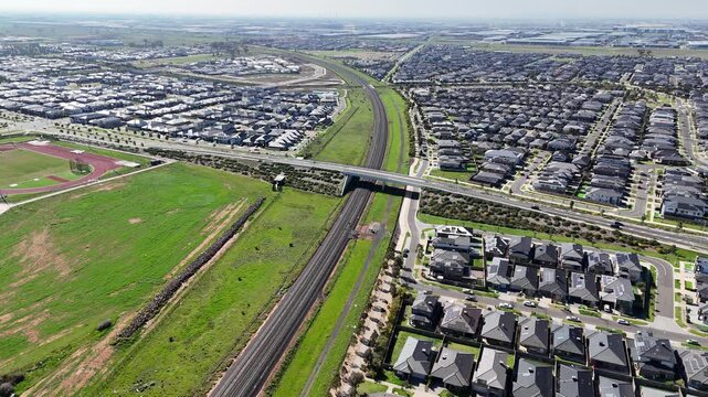 Aerial view of modern outer-suburban housing estate featuring uniform grey tiled roofs, structured street grids, and the regional railway line in Truganina, Melbourne, Victoria, Australia.