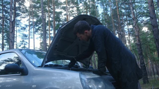 Young man in work clothes trying to repair a broken down car with an open hood in a forest, checking the engine with a screwdriver and experiencing difficulties with the vehicle maintenance