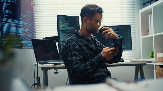Young Caucasian male software engineer enjoy coffee break with smartphone during work at desk. Workday productivity and concentration, Freelance career lifestyle, Modern tech office workplace concept.