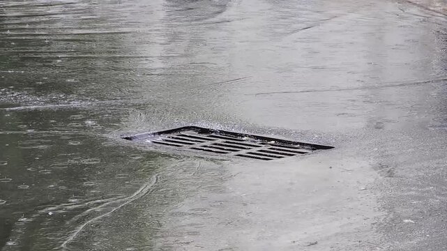 Storm drain on wet asphalt surface during heavy rain shower water flowing into drain grating creating ripples and reflections of environment on damp pavement