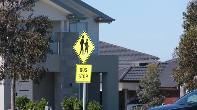 A yellow pedestrian crossing warning sign and bus stop sign in front of modern suburban homes beside a road with busy traffic, to alert drivers to slow down.  Melbourne, Victoria, Australia. 