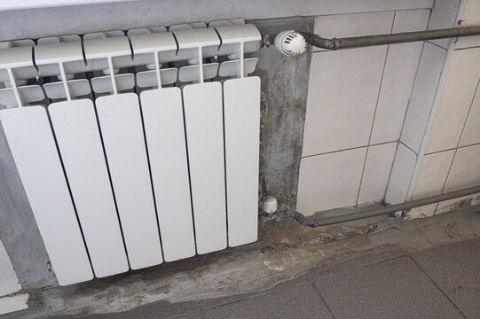 white bimetallic radiator mounted under tiled wall, connected to metal pipes. visible wear on wall and floor reveals an aged utility space