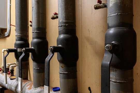 Industrial heating pipes with black expansion tanks, metal brackets, brass valves against wooden wall. visible welds, bolts, aged metal surfaces