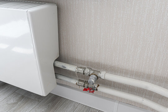 white radiator with bottom connection features metal-plastic pipes and red shut-off valve. textured beige wall and light-wood flooring complete the utility setup